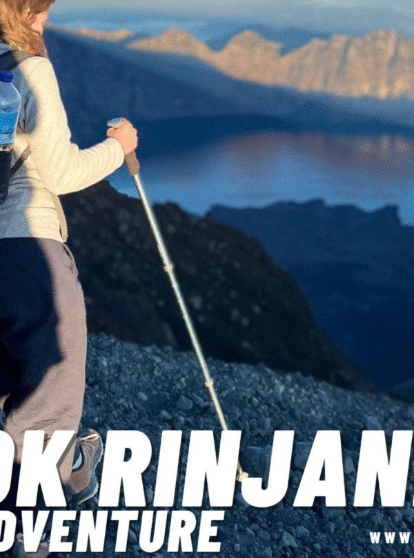 Female hiker with a backpack and trekking poles traversing a rocky slope on Mount Rinjani, overlooking the vast caldera and Segara Anak lake below. The LRM logo is visible in the corner.