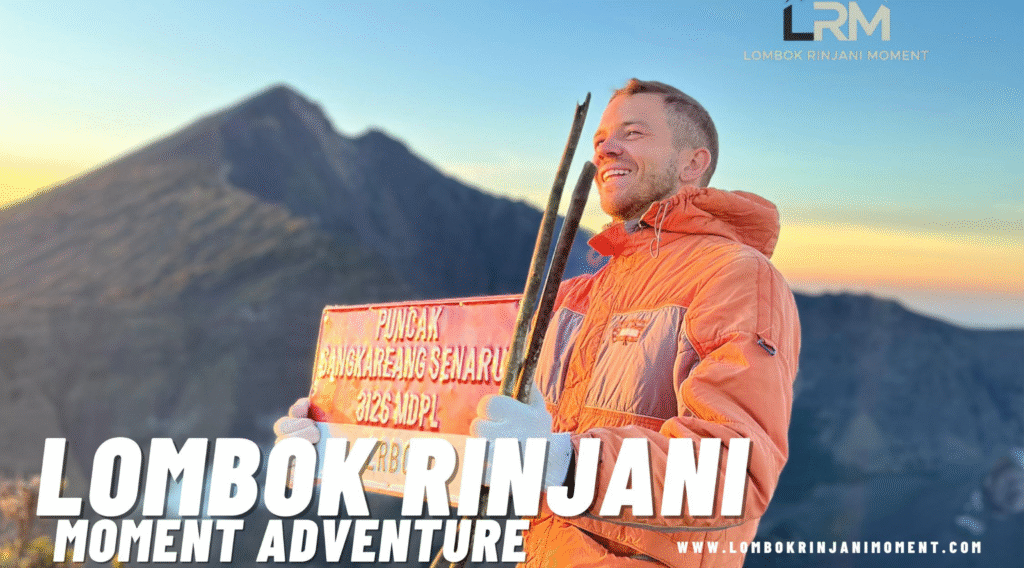 A smiling male hiker holding a red sign for Puncak Sangkareang Senaru (3125 Mdpl) on Mount Rinjani at sunrise, wearing an orange jacket, with the volcano peak in the background.
