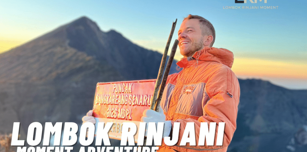 A smiling male hiker holding a red sign for Puncak Sangkareang Senaru (3125 Mdpl) on Mount Rinjani at sunrise, wearing an orange jacket, with the volcano peak in the background.