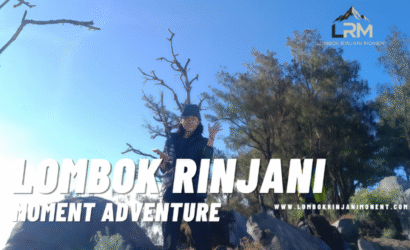 A female hiker wearing a cap and jacket poses with hands up against a bright blue sky, framed by dry, dark tree branches and green trees on the Rinjani trail.
