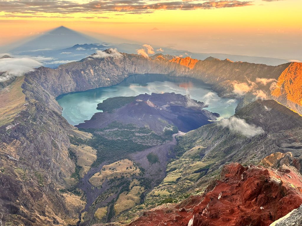 A beautiful view from the summit of Mount Rinjani, showcasing the vast crater and Segara Anak lake below, surrounded by mountains and a clear blue sky.