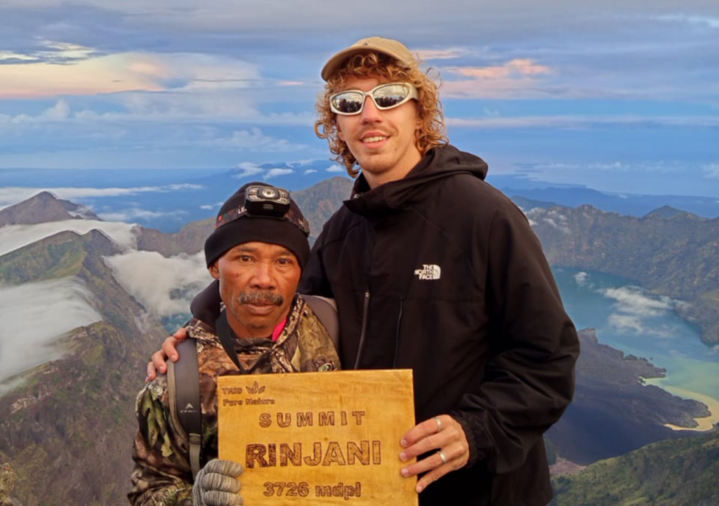 A close-up portrait of a foreign male hiker with blonde hair and sunglasses standing closely alongside his local mountain guide Yasin at the Mount Rinjani summit, proudly holding a wooden 3,726 mdpl sign with the crater lake in the background. www.lombokrinjanimoment.com