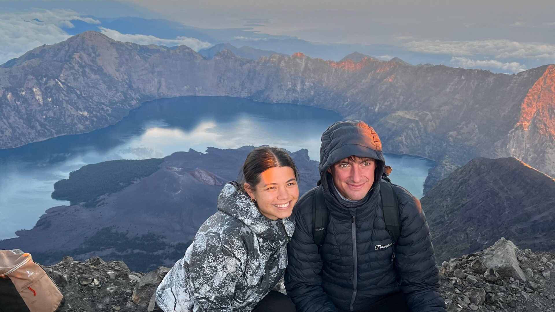 A pair of hikers smiling at the summit of Mount Rinjani in Lombok, Indonesia. They are holding a sign that reads 3726 mdpl, with the caldera of Lake Segara Anak and a sunrise in the background. They are wearing thick jackets and pants, looking happy after reaching the peak.